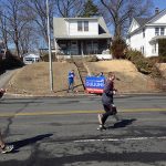 A person runs down a street as people in the background hold a sign for Hampden District Attorney Anthony Gulluni.