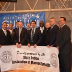 Hampden District Attorney Anthony Gulluni poses for a photo with people in suits as they hold a sign that reads "Proudly endorsed by the state police association of Massachusetts."