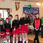 A group of girls wearing identical red skirts and black blouses and a boy wearing a red tie and red vest stand and listen.