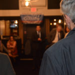 A blurry photo of an event with a man facing the opposite direction in the foreground